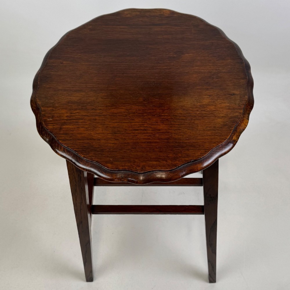 A tall, dark oak Edwardian side table featuring a scalloped piecrust rim top, four splayed square legs, and a lower slatted shelf. View VII