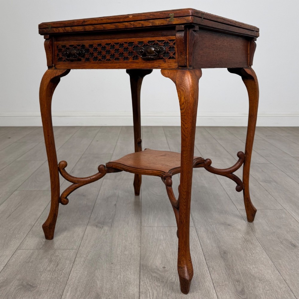 An Edwardian oak envelope card table featuring cabriole legs, a decorative carved drawer front, and a shaped lower shelf. View II