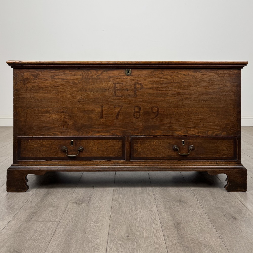 An 18th-century Georgian golden oak mule chest featuring a hinged lid, two lower drawers with brass drop handles, shaped bracket feet, and the inscribed date 1789.