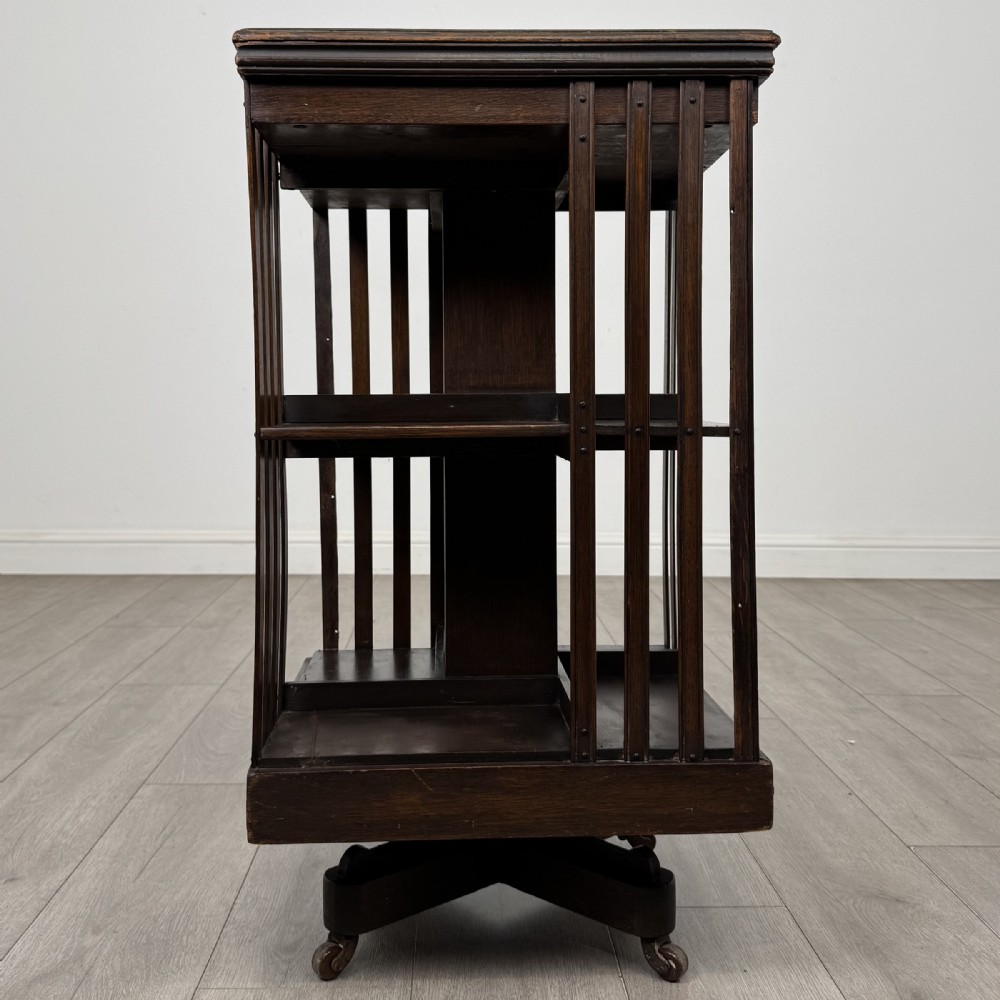 A dark oak Edwardian revolving bookcase with vertical slatted sides, two shelf tiers, and a quadform base on castors.