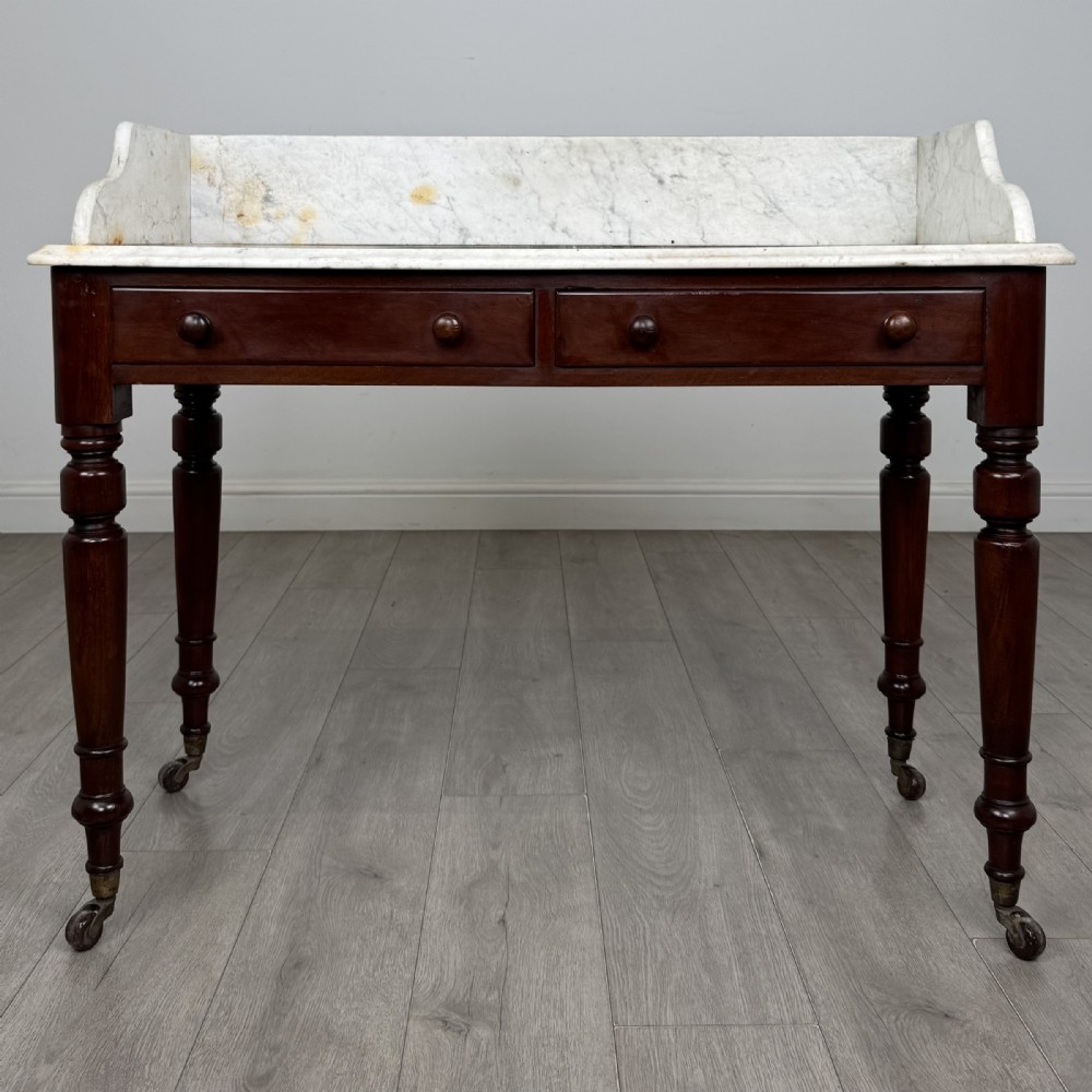 A Victorian mahogany washstand featuring a white marble top with a raised splashback, two drawers with turned knobs, turned legs, and brass castors.