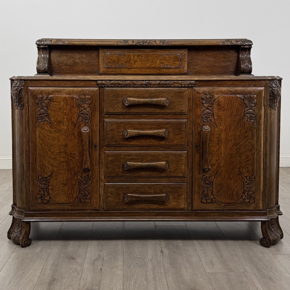 An antique golden oak Art Deco sideboard featuring a carved gallery back, four central drawers, flanked cupboard doors with carved floral panels, and sculpted wooden handles.