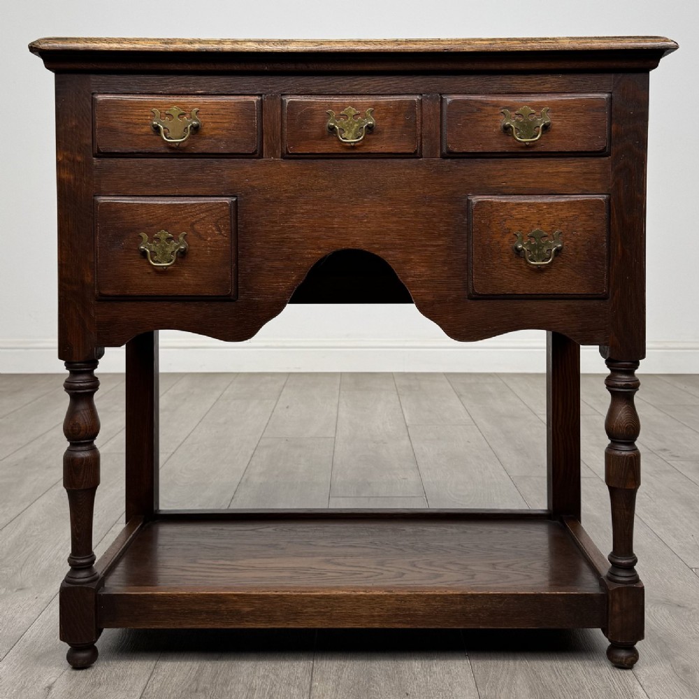 A dark oak side table featuring five drawers with brass handles, turned front legs, an arched kneehole, and a lower tier shelf.