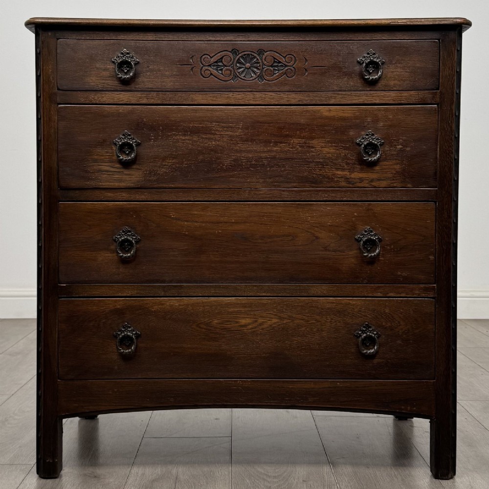 A dark brown carved oak chest of drawers featuring four stacked drawers with drop ring handles and an incised rosette carving on the top drawer.