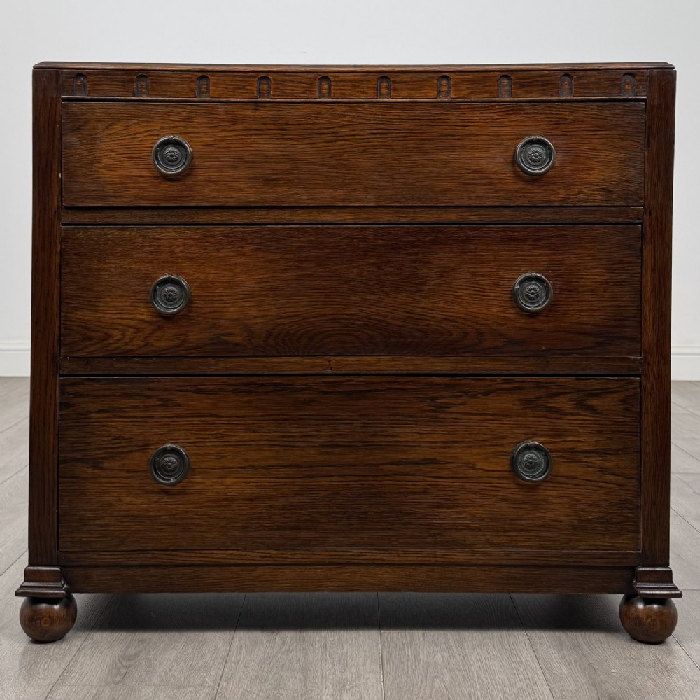 A dark oak chest of three drawers featuring carved decorative detailing along the top edge, circular ring handles, and turned bun feet.