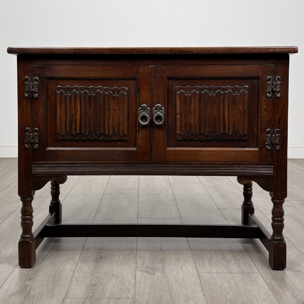 A dark oak cupboard featuring two linenfold carved door panels, decorative metal hinges, ring pull handles, turned legs, and a lift top.
