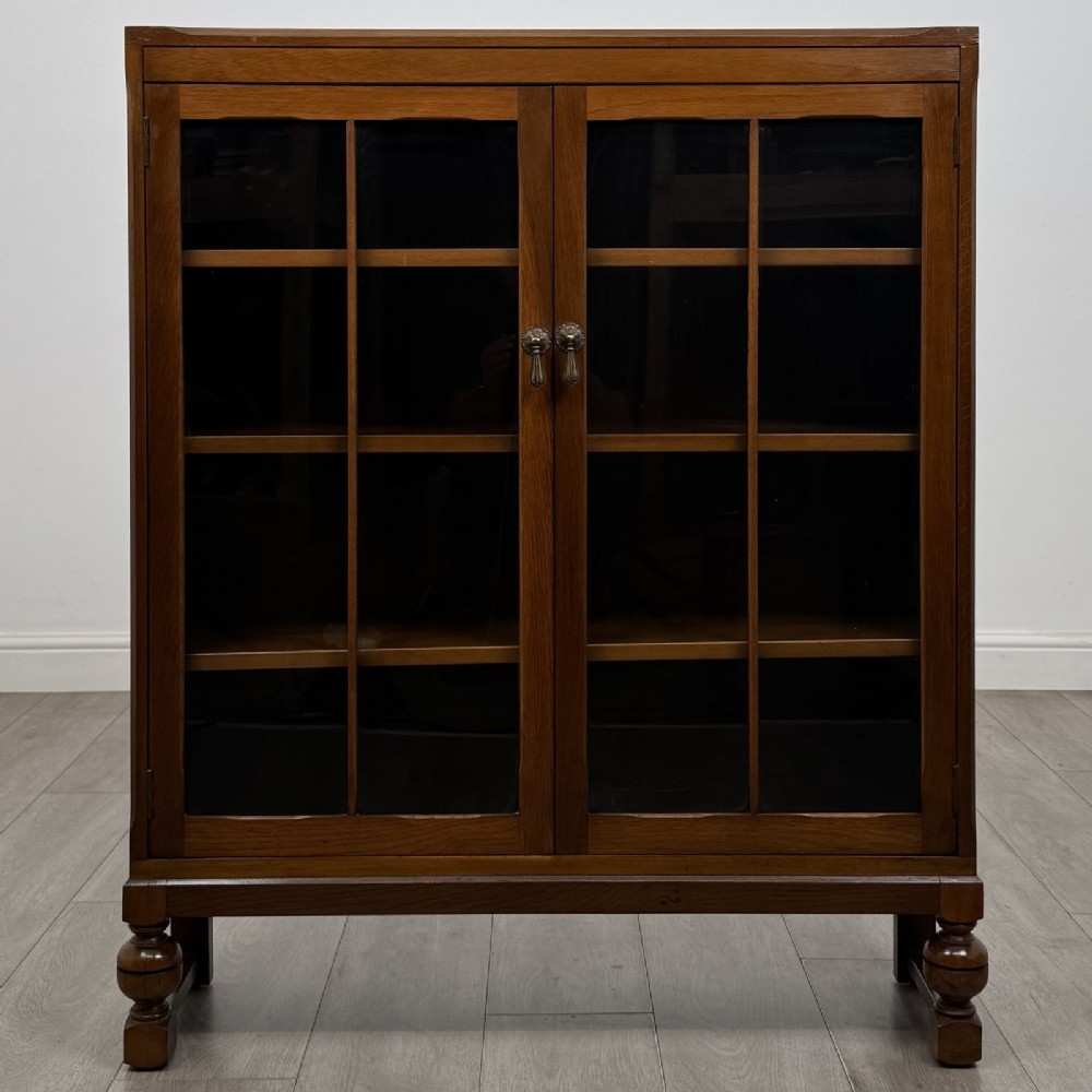 A warm brown antique oak display cabinet featuring two glazed doors with wooden glazing bars and squat turned legs.