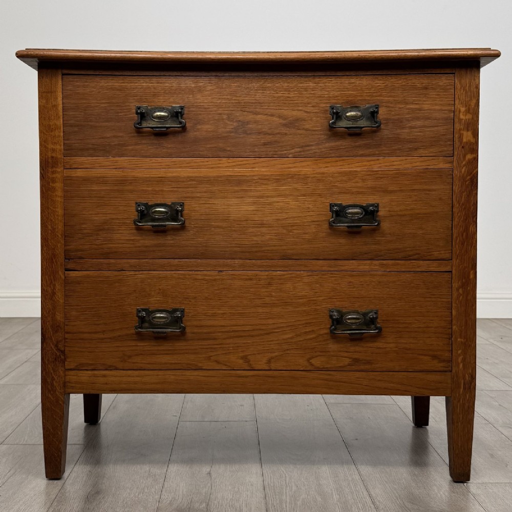 An Edwardian golden oak chest of drawers featuring three full-width drawers with aged brass drop handles, supported by square tapered legs.