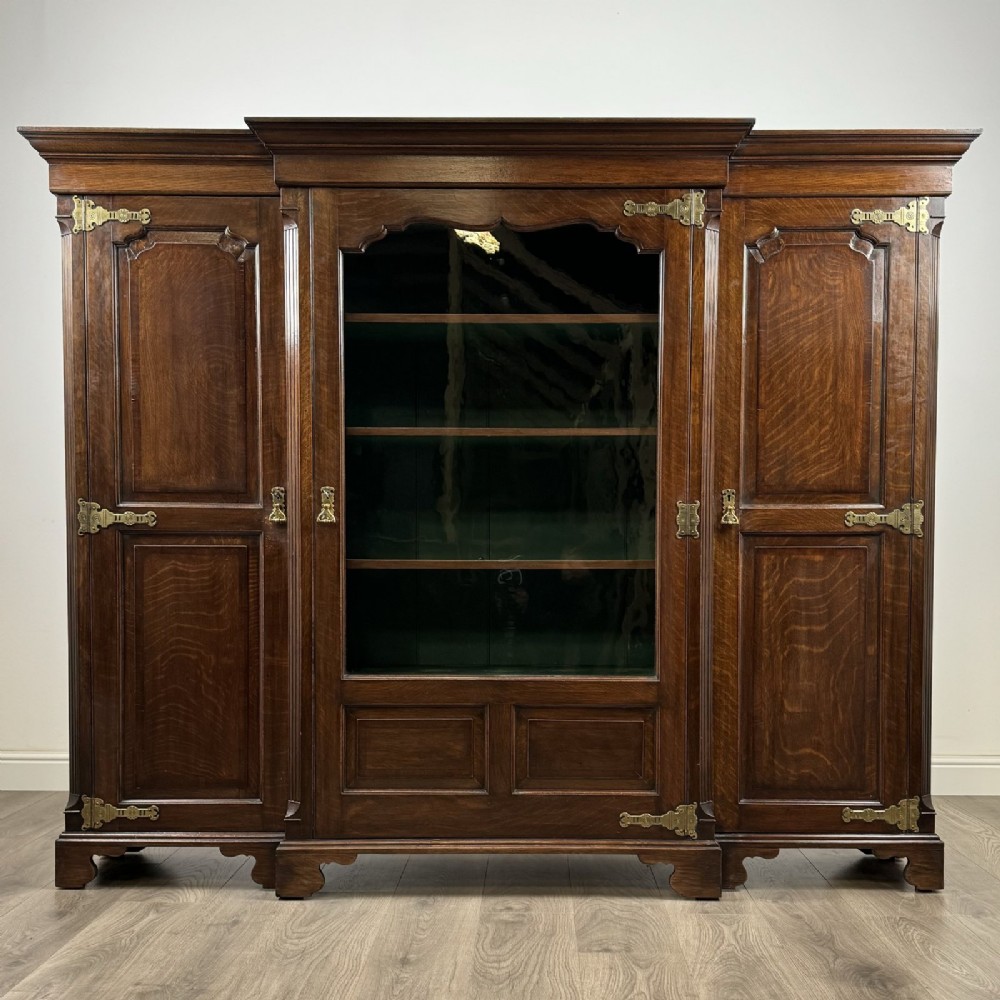 A large dark oak display cabinet featuring a central glazed door with shelves, flanked by two solid panelled doors adorned with decorative brass strap hinges.