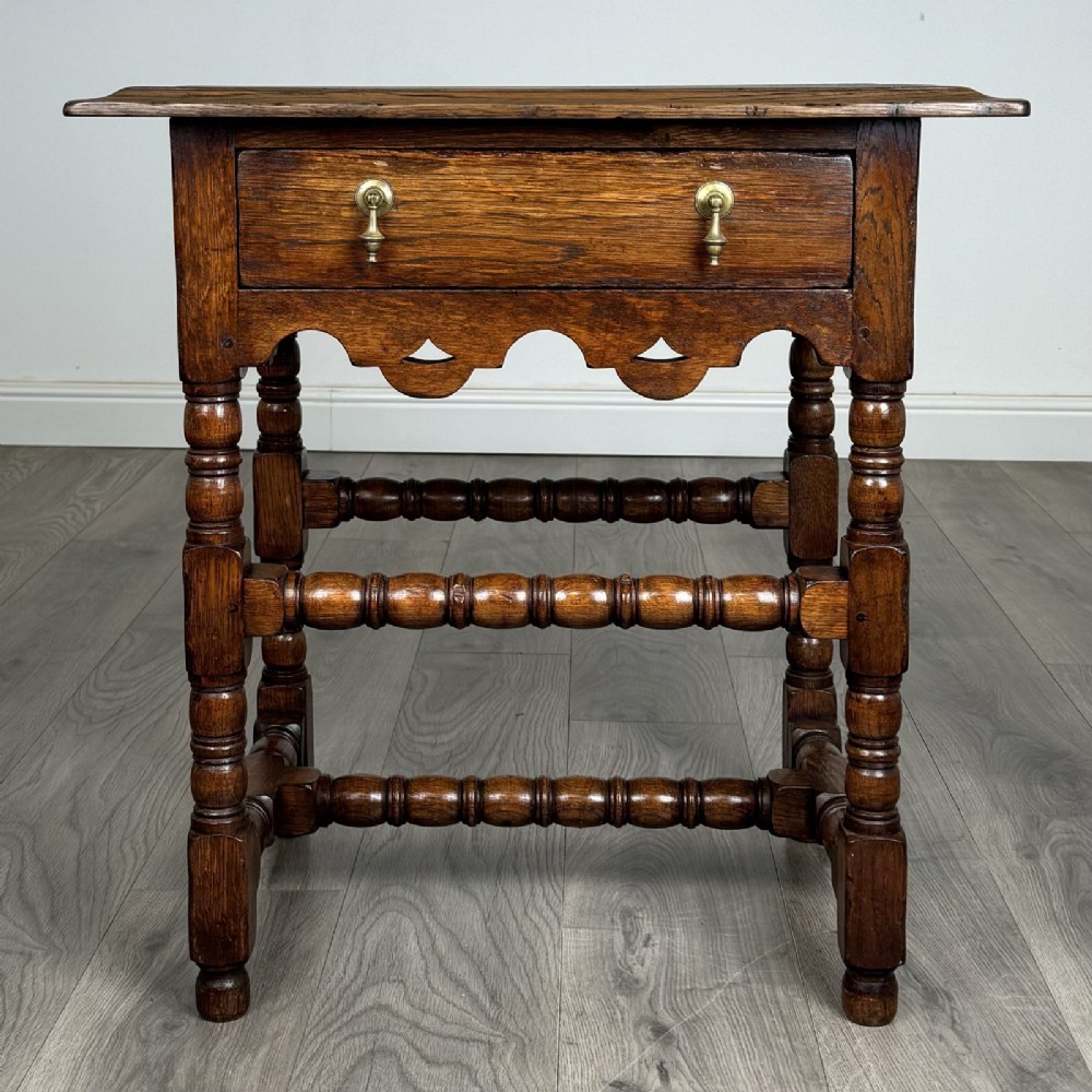 An antique dark oak side table featuring a single drawer with brass drop handles, a scalloped apron, and bobbin-turned legs connected by matching stretchers.