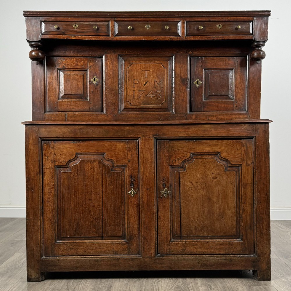 An 18th-century Georgian dark oak Welsh deuddarn cupboard featuring carved upper panels dated 1743, turned drop pendants, and panelled cupboard doors.