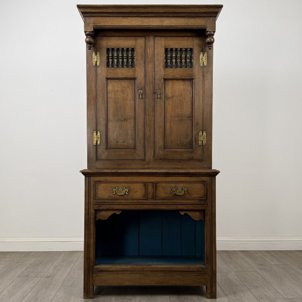 A tall antique brown oak cupboard with carved spindle panels on the upper cabinet doors, brass butterfly hinges, two drawers, and a lower open alcove with a blue painted interior.
