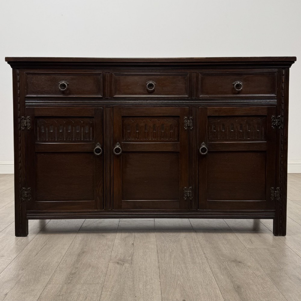 A dark oak sideboard featuring three top drawers with metal ring pulls above three carved panel cupboard doors.