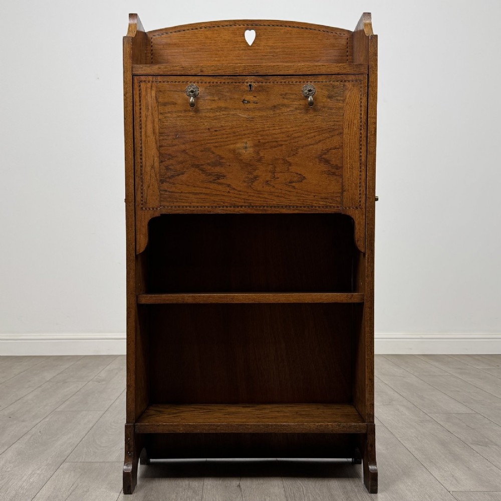 An Arts and Crafts golden oak bureau with a heart cutout on the top gallery back, a fall-front writing surface with drop handles, and two open bookshelves underneath.