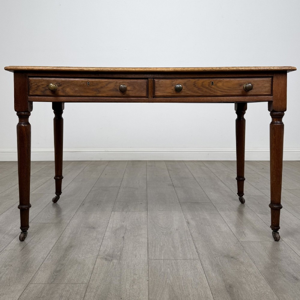 An antique golden oak writing table featuring two frieze drawers with turned knobs and supported by turned legs on brass castors.