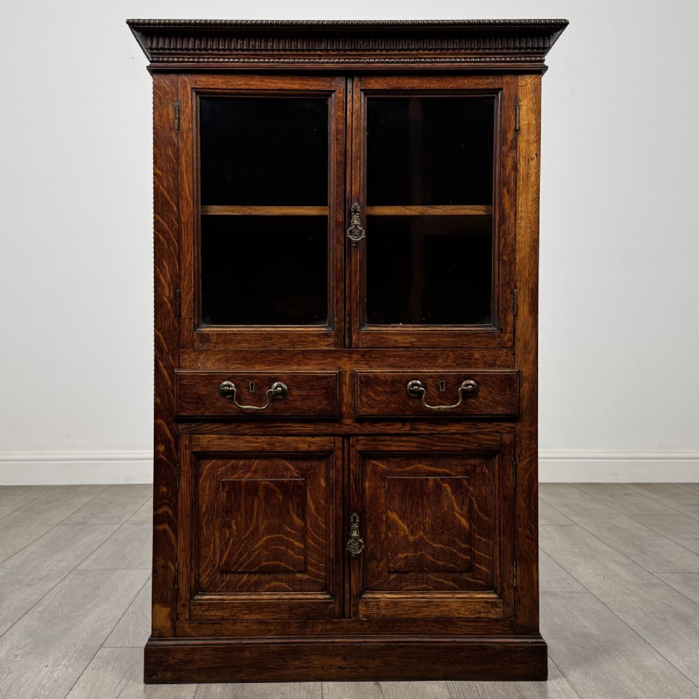 A dark oak Victorian display cabinet featuring a dentil cornice, glazed upper doors, two drawers with brass handles, and panelled lower cupboard doors.