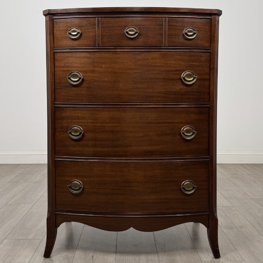 An antique mahogany chest of drawers with a bowfront shape, brass oval handles, and reeded side columns.