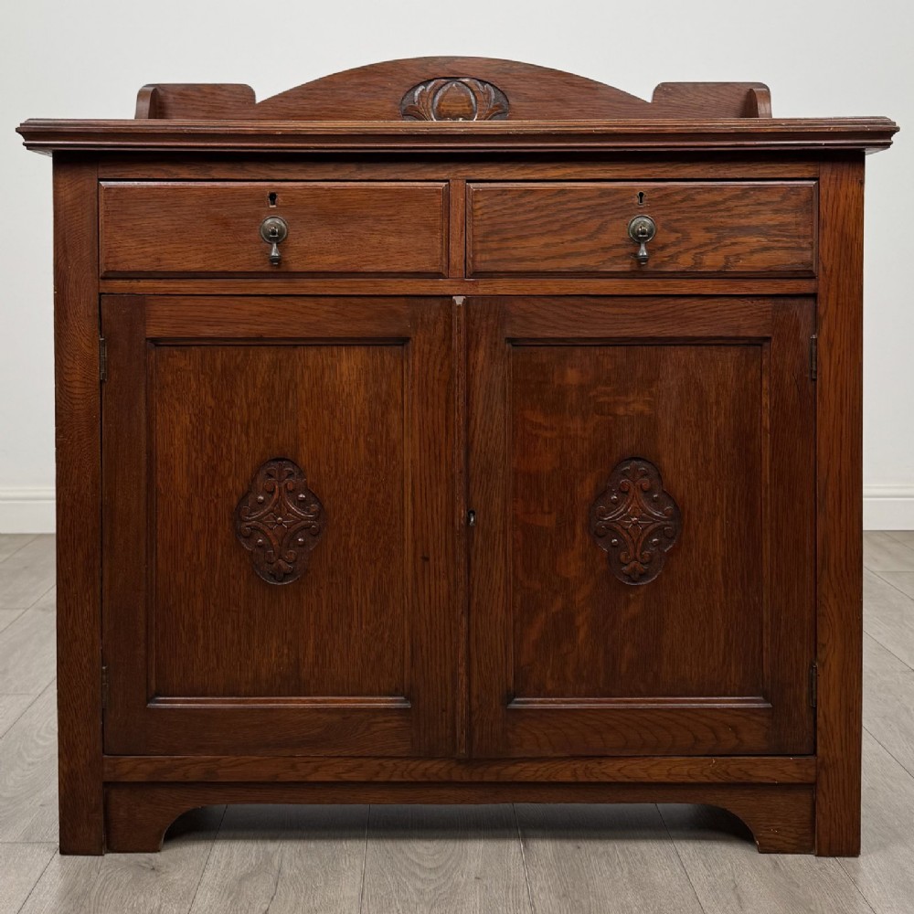 An Edwardian dark oak cupboard featuring a raised carved gallery, two top drawers with brass drop handles, and two panelled doors decorated with carved central cartouches.