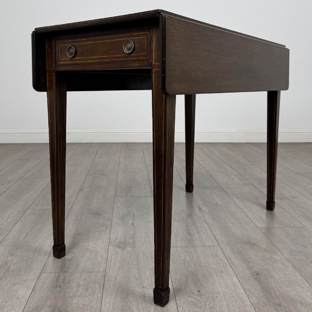A dark brown mahogany Pembroke table featuring two drop flaps, a front drawer with boxwood line inlay, two brass ring handles, and square tapered legs ending in spade feet.