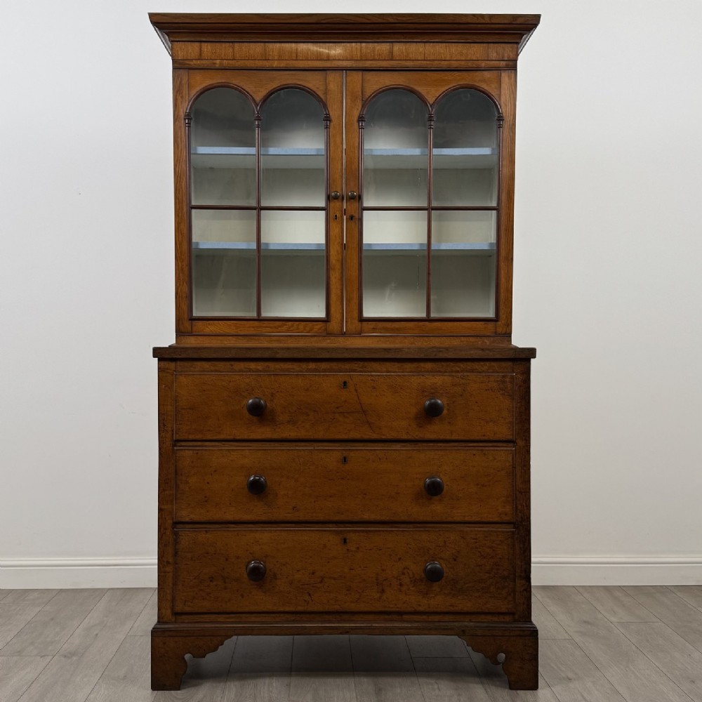 An 18th-century Georgian golden oak glazed bookcase resting upon a chest of three long drawers with turned wooden handles and shaped bracket feet.