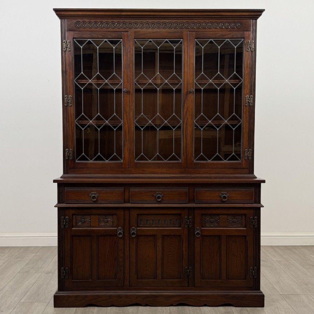 A dark oak display cabinet featuring three leaded glass doors, three drawers, and three carved panelled cupboards with brass handles.