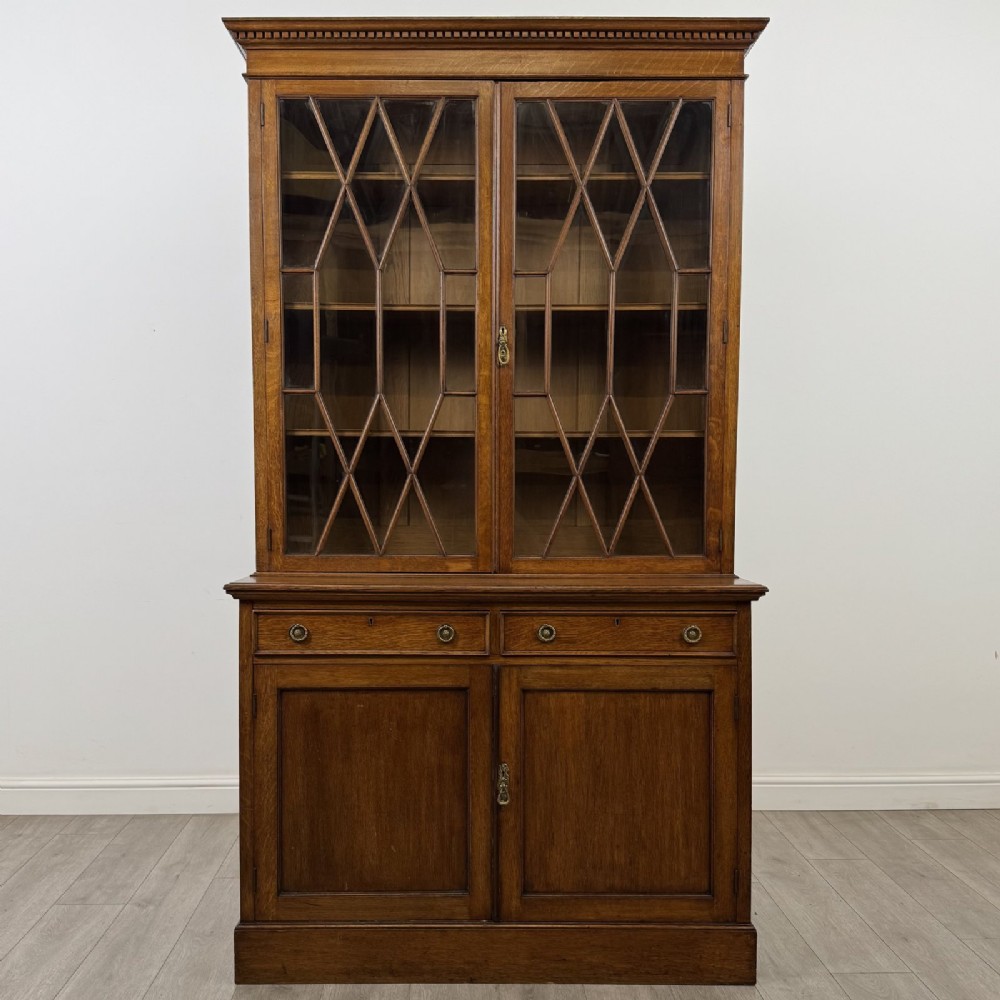 A Victorian golden oak two-part glazed bookcase featuring geometric astragal glazing, a dentil cornice, two drawers, and a panelled base cupboard.