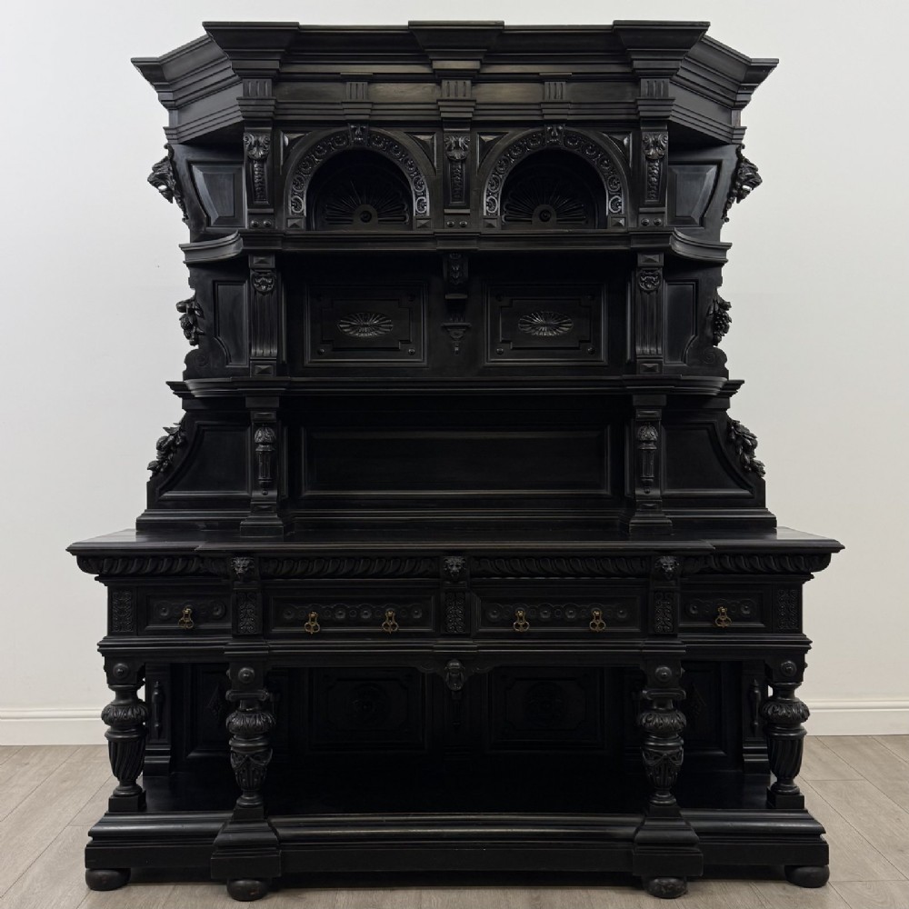 An ornate ebonised dark wood Victorian sideboard featuring upper tier shelves with carved arches, lion masks, and lower drawers with brass drop handles.