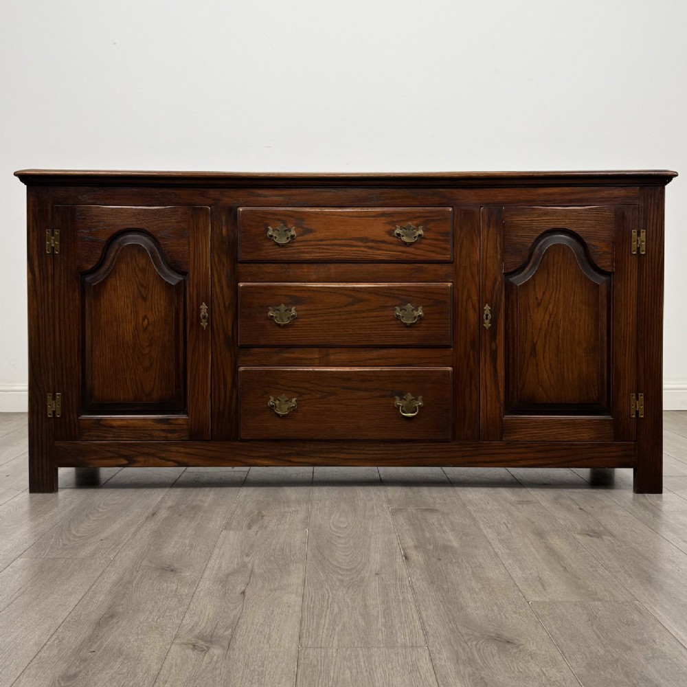 A dark oak sideboard featuring three central drawers flanked by two arched panel cupboard doors with brass handles.