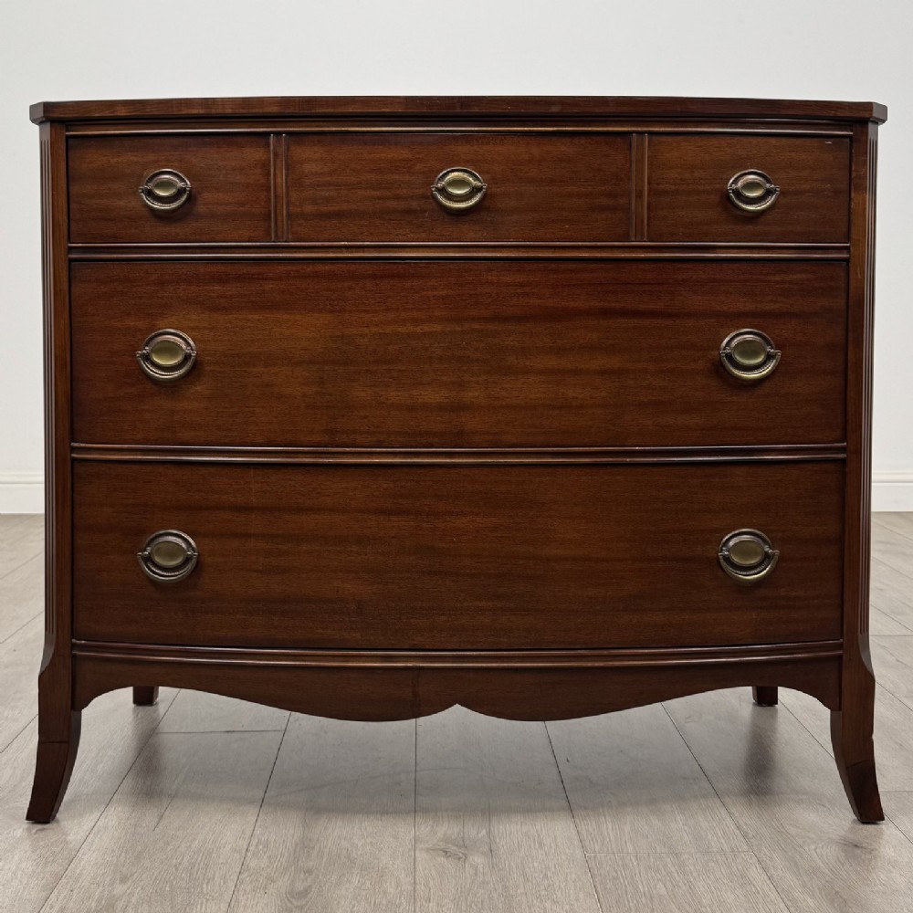 A dark mahogany bow-fronted chest of drawers featuring brass oval ring handles, reeded side columns, and curved legs.