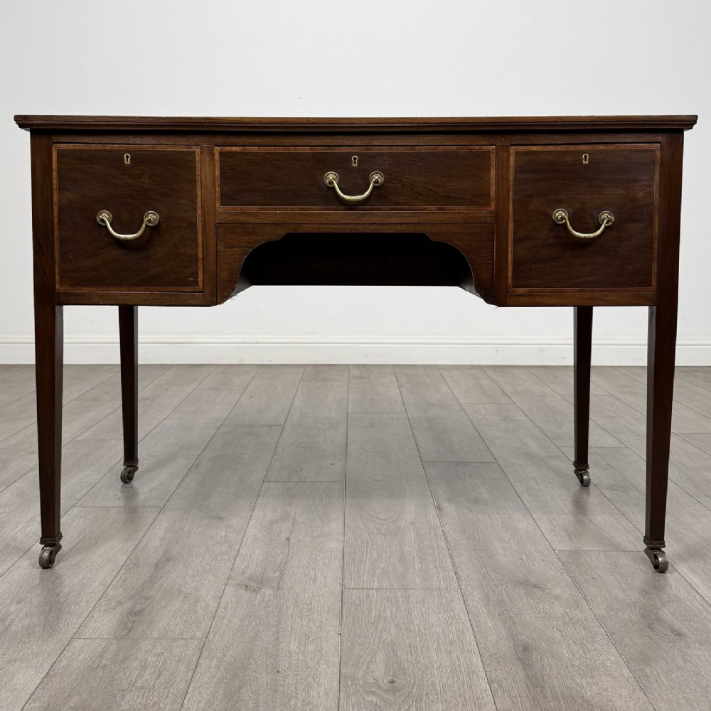 An Edwardian mahogany writing table featuring three inlaid drawers, brass drop handles, square tapered legs, and brass castors.
