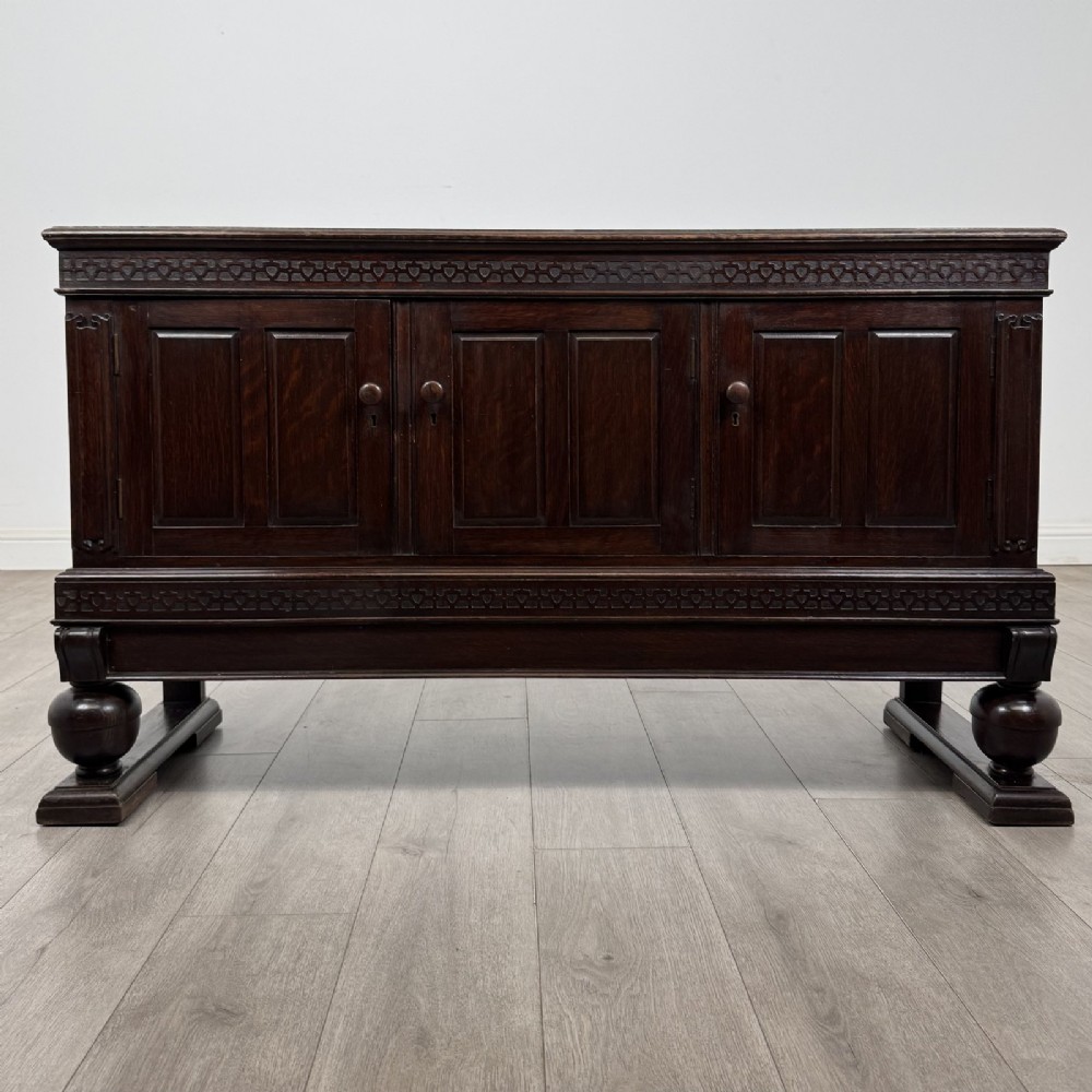A dark oak Edwardian low cupboard featuring three panelled doors, carved frieze borders, and turned bulbous legs on sledge feet.