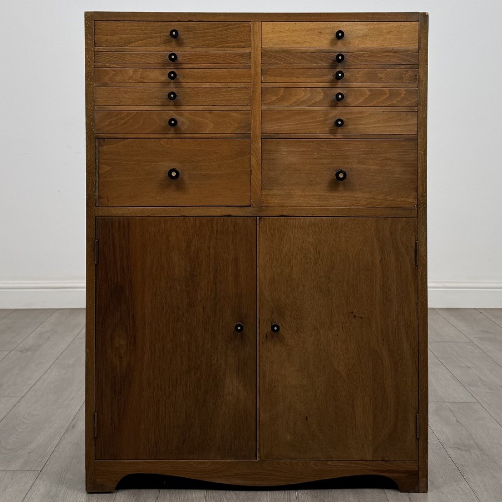 An antique oak storage cabinet featuring multiple shallow drawers arranged above a double-door lower cupboard with small turned dark wooden knobs.
