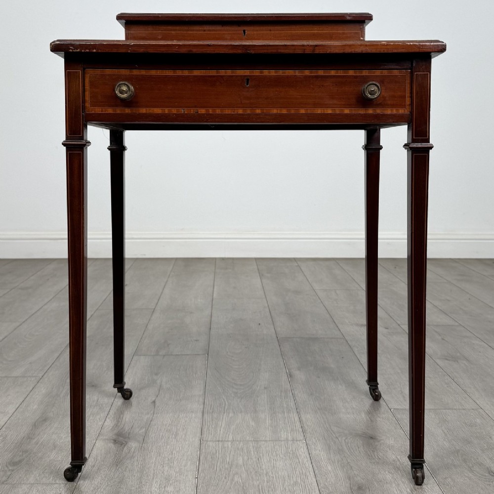 An Edwardian mahogany writing table featuring boxwood inlay, a single drawer with brass knobs, a raised rear stationery shelf, and square tapered legs on castors.