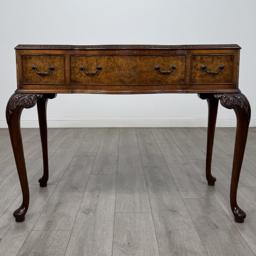 An antique burr walnut writing table featuring three drawers, carved cabriole legs, and decorative brass handles.