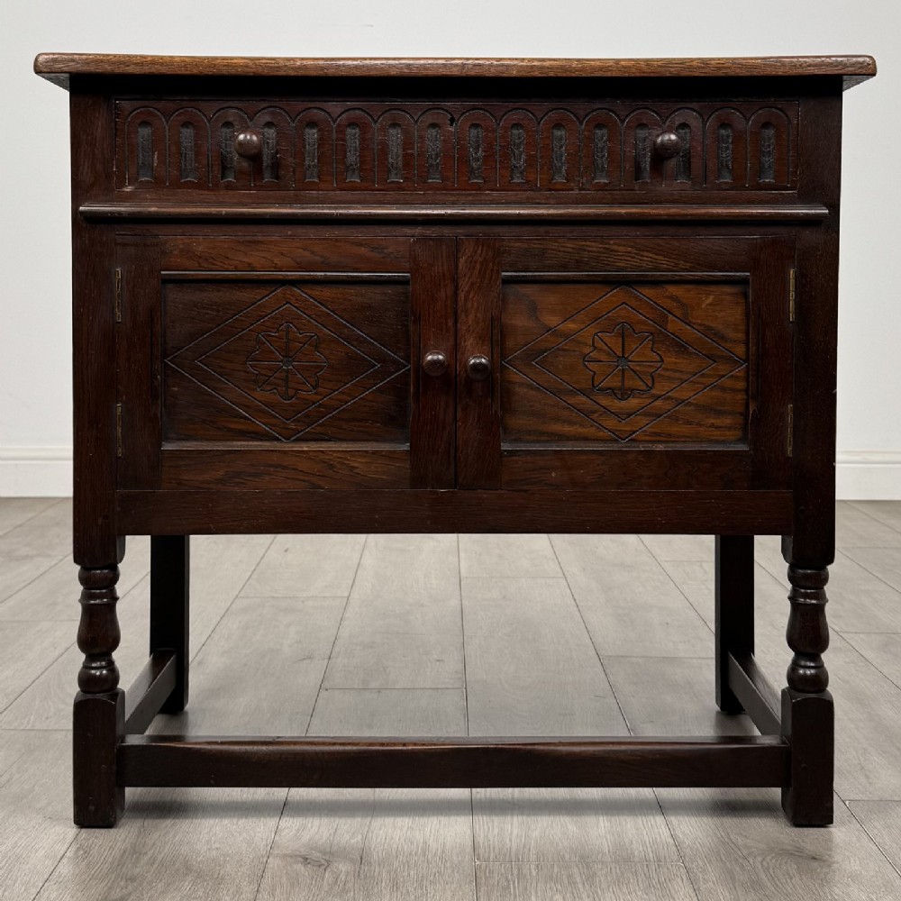 A dark brown oak antique cupboard featuring a carved top drawer, panelled double doors with carved diamond motifs, and turned legs with stretchers.