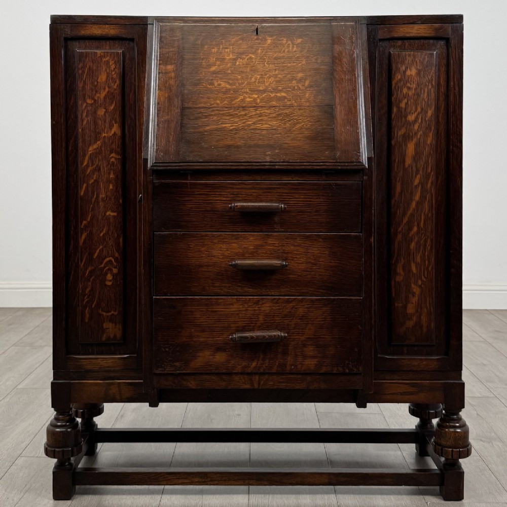 A dark oak fitted bureau featuring a central slant-front writing slope over three drawers, flanked by two side cupboard doors and supported on turned feet with lower stretchers.