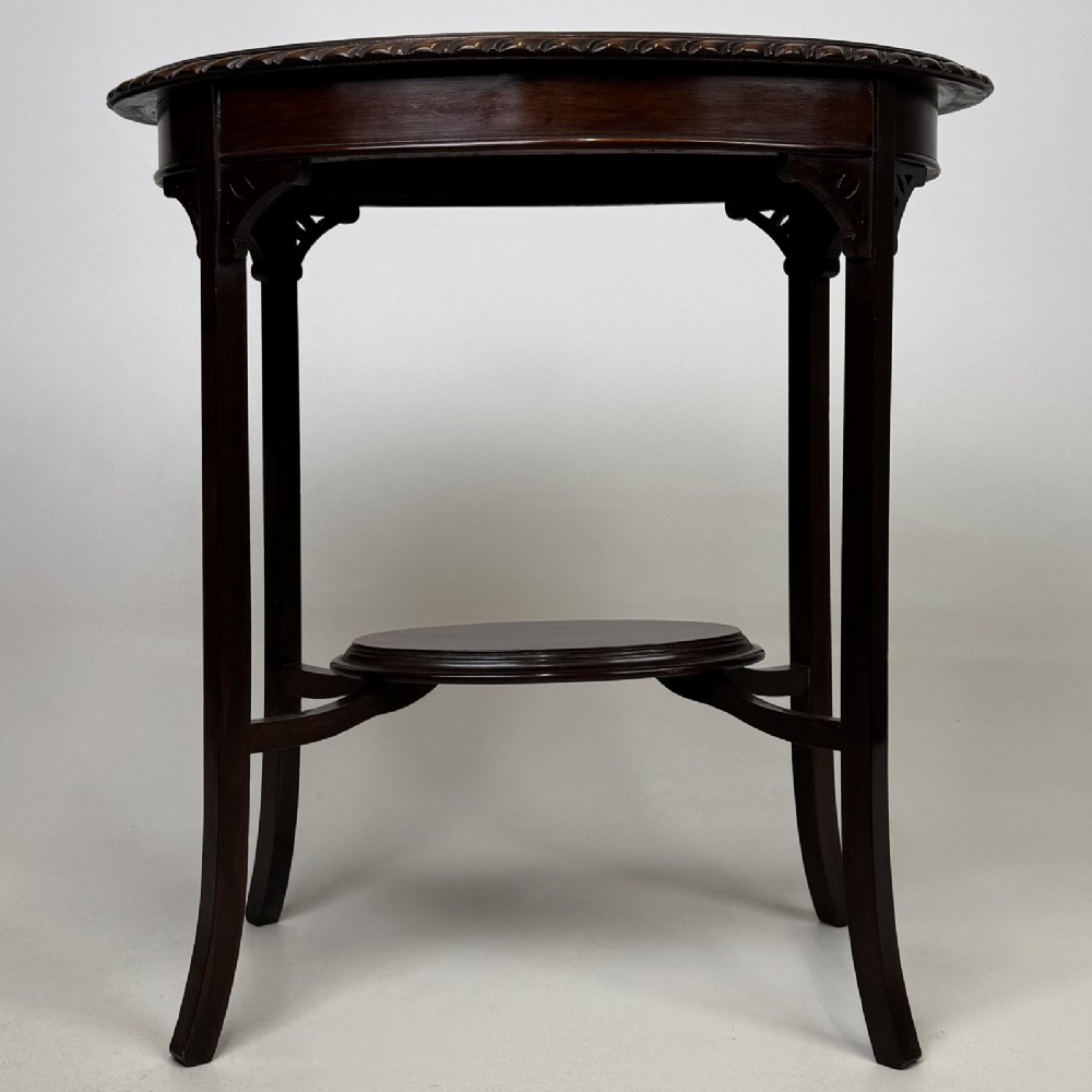 An Edwardian oval mahogany side table featuring a carved gadrooned top edge, pierced corner brackets, slender tapered legs, and a lower oval shelf.