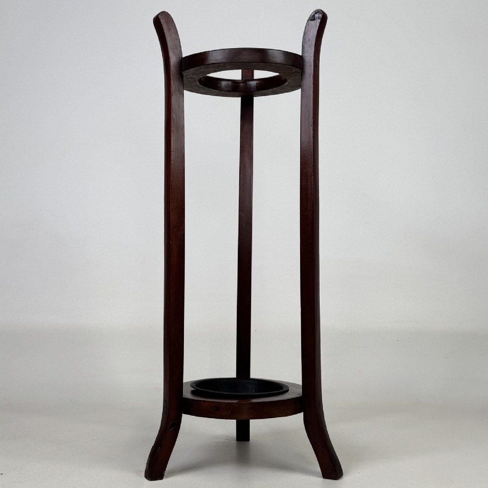 A tall, dark mahogany three-legged umbrella stand featuring a circular top ring and a metal drip tray built into the lower ring base.