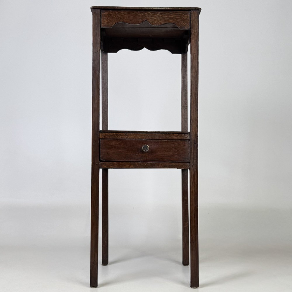 A tall dark oak Edwardian side table featuring a top surface with a scalloped apron shelf beneath, a single drawer with a central brass knob, and four slender square legs.