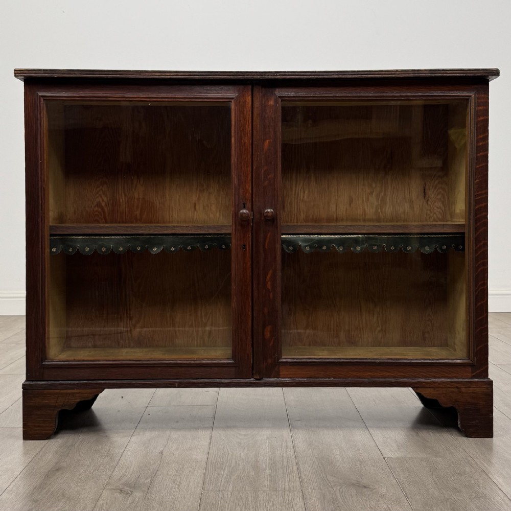 A low Victorian dark oak display cabinet featuring two glazed doors, turned wooden knobs, bracket feet, and a single interior shelf trimmed with scalloped leather.