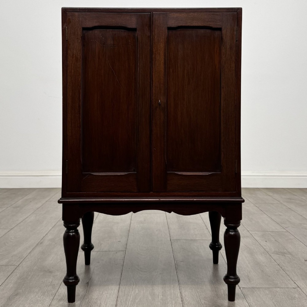 A small dark mahogany cabinet featuring two panelled doors, a shaped apron, and four turned legs.