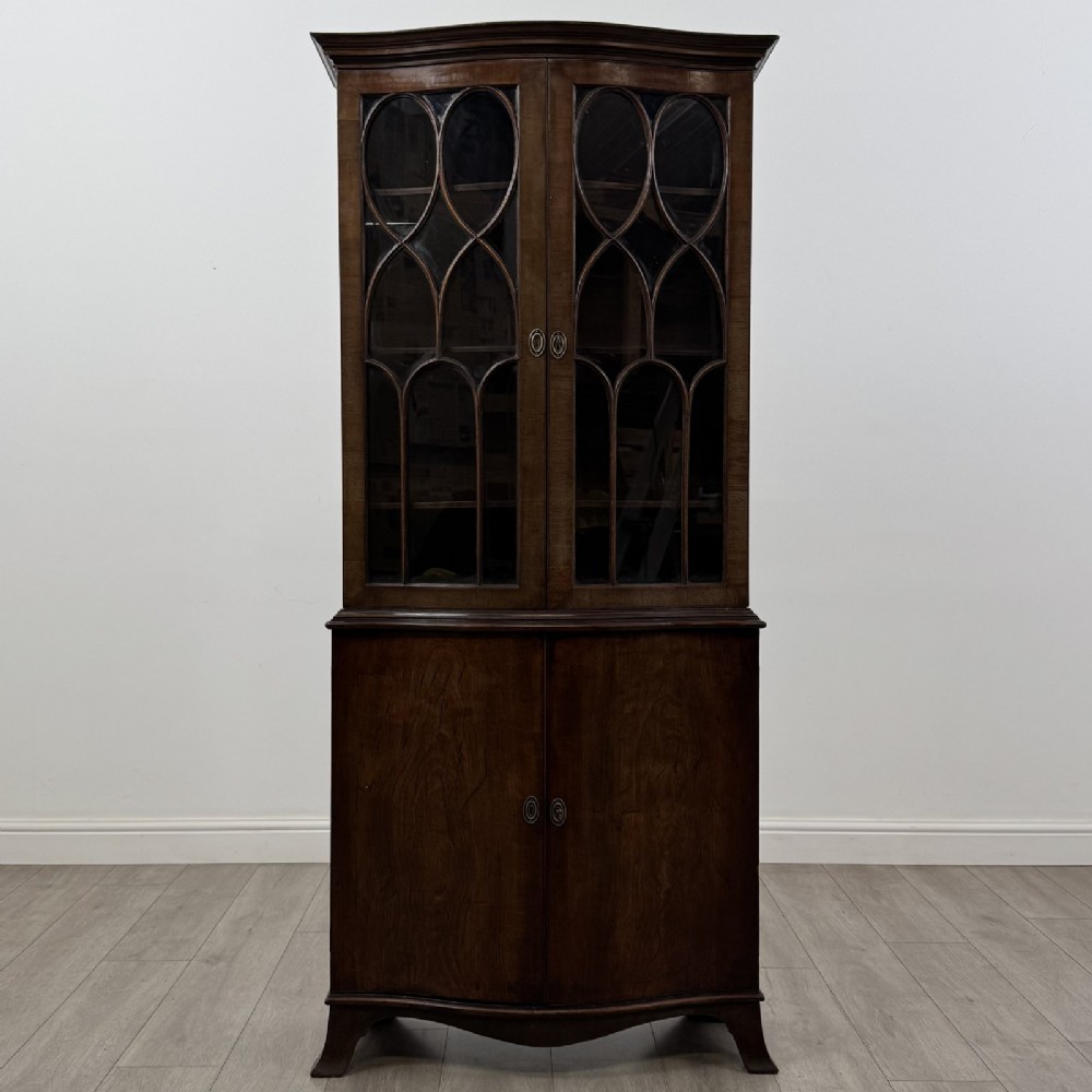 A tall dark mahogany bookcase featuring a serpentine front profile, upper astragal glazed double doors, and a lower double-door cupboard.