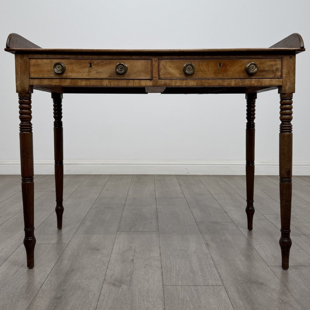 An antique Georgian mahogany writing table featuring a raised gallery edge, two frieze drawers with brass circular handles, and slender turned legs.