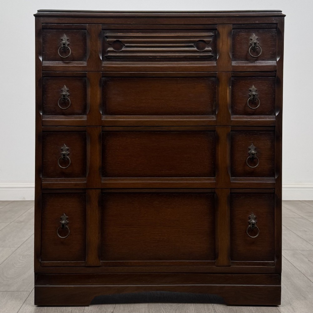 A dark brown oak chest of drawers with geometric panel moulding, four horizontal drawers, ornate metal ring handles, and a plinth base.