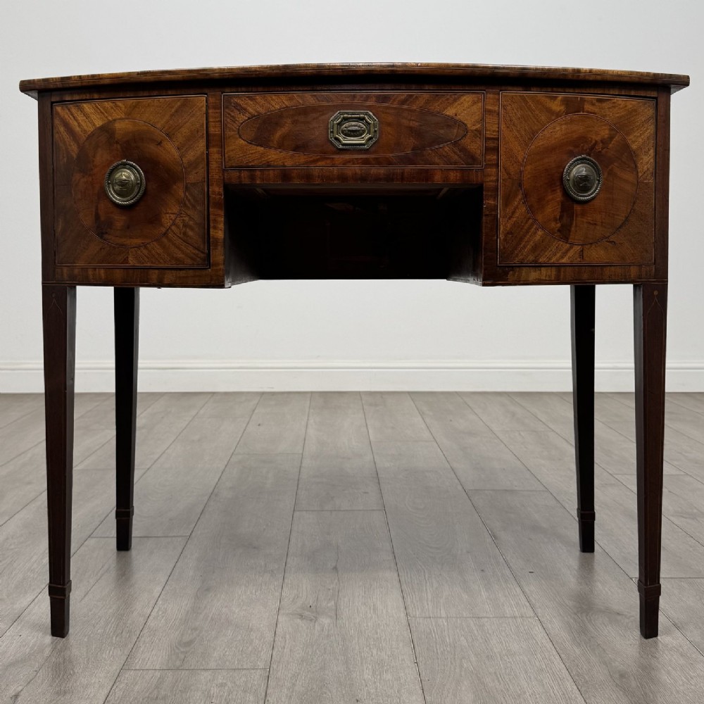 An antique Georgian mahogany bow-front side table with flame mahogany inlaid drawers, brass ring handles, and tapered square legs.