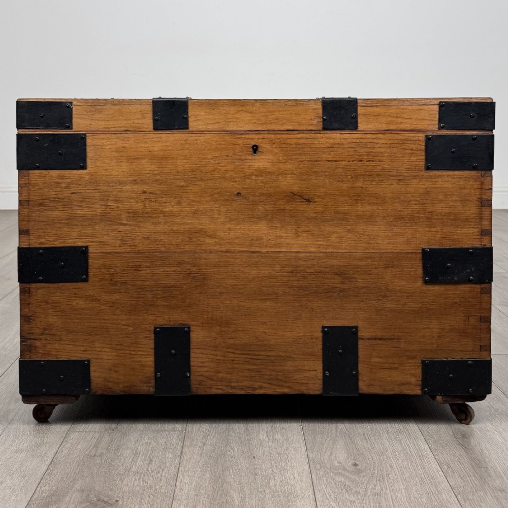 A rectangular warm brown oak storage chest featuring black metal strap reinforcement, a keyhole, and corner brackets on small castors.