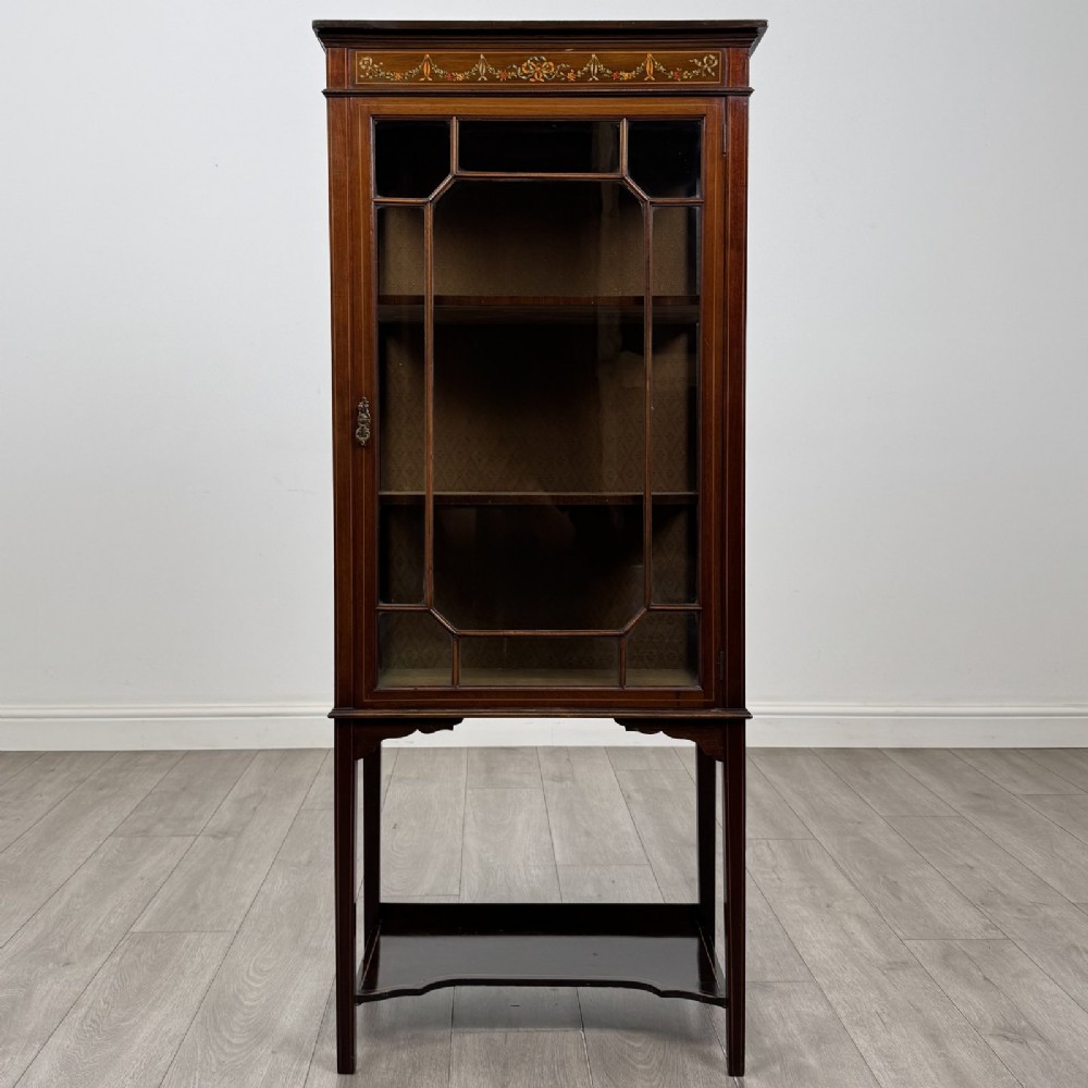 An Edwardian mahogany display cabinet featuring a painted inlaid floral frieze, single astragal glazed door, fabric-lined interior shelves, and tapered legs with a lower undertier.