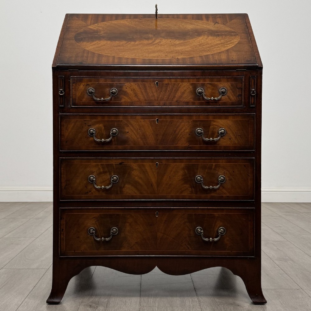 A small Georgian style dark mahogany writing bureau featuring an inlaid oval motif on the slant-front lid and four drawers with brass handles.