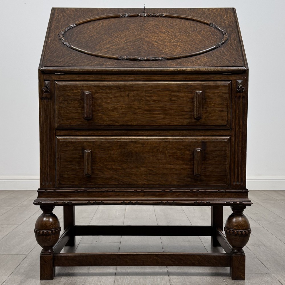 An antique dark oak fall-front writing bureau featuring a carved oval motif on the sloping lid, two lower drawers with wooden handles, and bulbous turned front legs on a stretcher base.