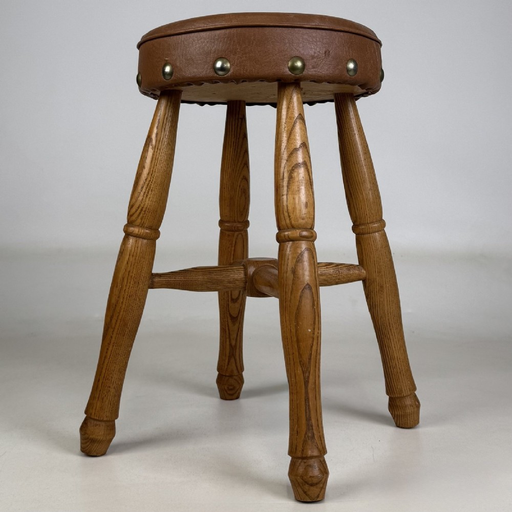 A circular antique oak stool featuring a padded brown leather seat secured with brass studs, supported by four turned legs and cross stretchers.